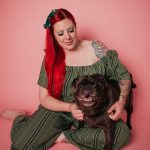 A full-length studio portrait of photographer Nadia De Sensi with vibrant red hair, smiling as she sits cross-legged against a solid pastel pink backdrop. She is wearing a green-and-dark-striped off-the-shoulder jumpsuit with a matching green bow in her hair, happily holding and posing with her dark black pug, Niles, who is looking directly at the camera with an open, happy expression