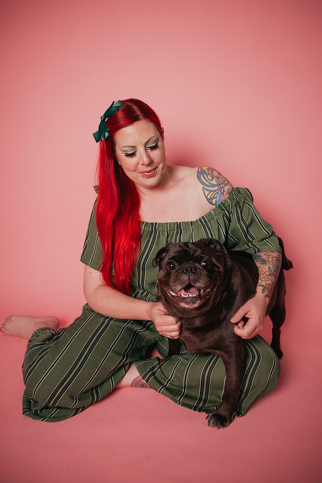 A full-length studio portrait of photographer Nadia De Sensi with vibrant red hair, smiling as she sits cross-legged against a solid pastel pink backdrop. She is wearing a green-and-dark-striped off-the-shoulder jumpsuit with a matching green bow in her hair, happily holding and posing with her dark black pug, Niles, who is looking directly at the camera with an open, happy expression