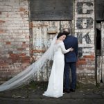 A romantic full-length wedding portrait of a bride and groom embracing outside on a paved path against a rustic brick wall and weathered wooden barn doors, captured by Nadia De Sensi.