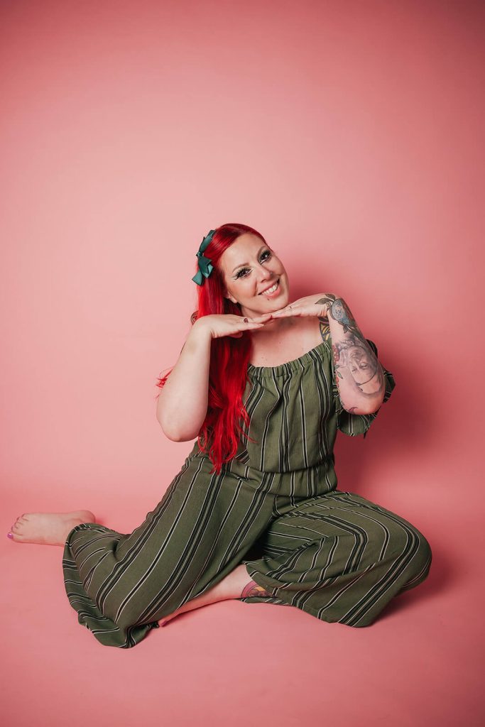 A vibrant, retro-conceptual portrait of Nadia De Sensi with a dark updo, bold winged eyeliner, and a red dress, smiling warmly against a pink studio background