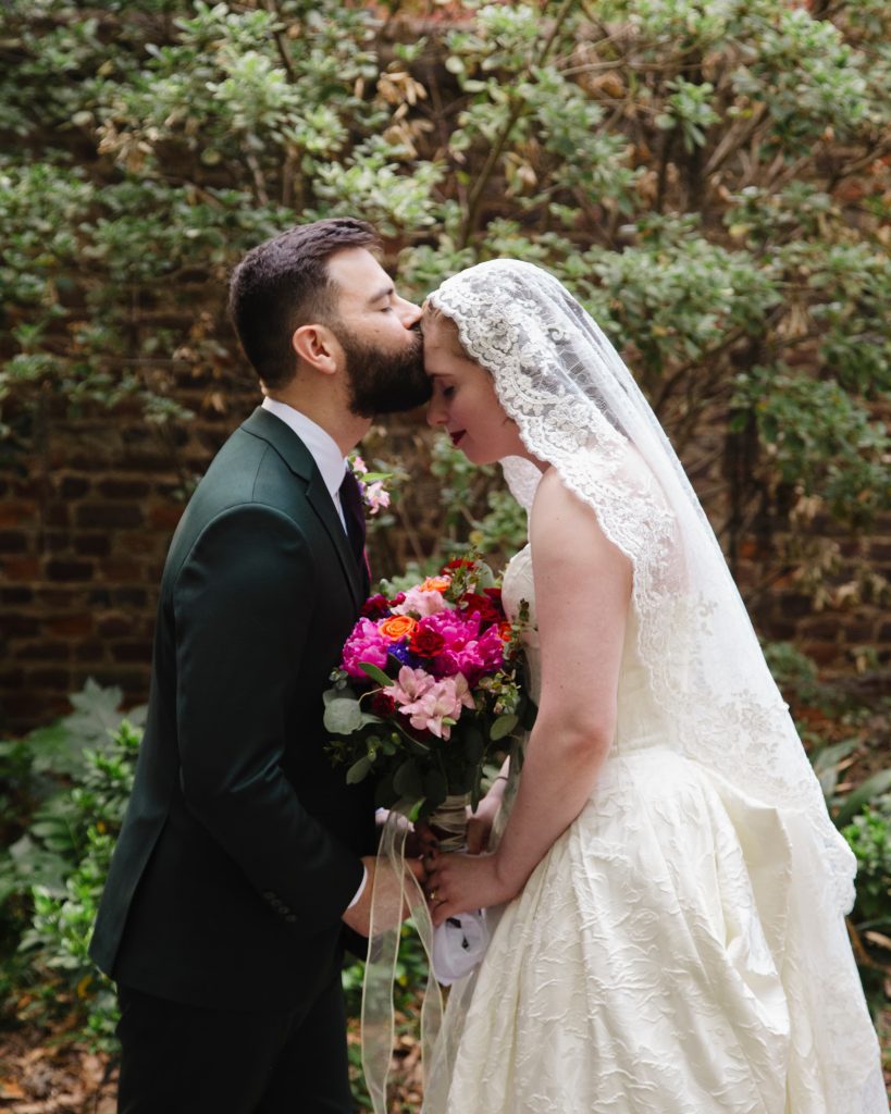 Bride wearing a lace wedding dress and holding a vibrant bouquet of bright pink, purple, and magenta flowers next to the groom in a dark suit.