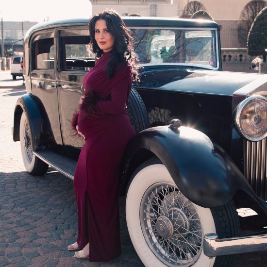 Pregnant woman in a burgundy dress posing beside a vintage dark blue car at Jefferson Hotel in Richmond VA