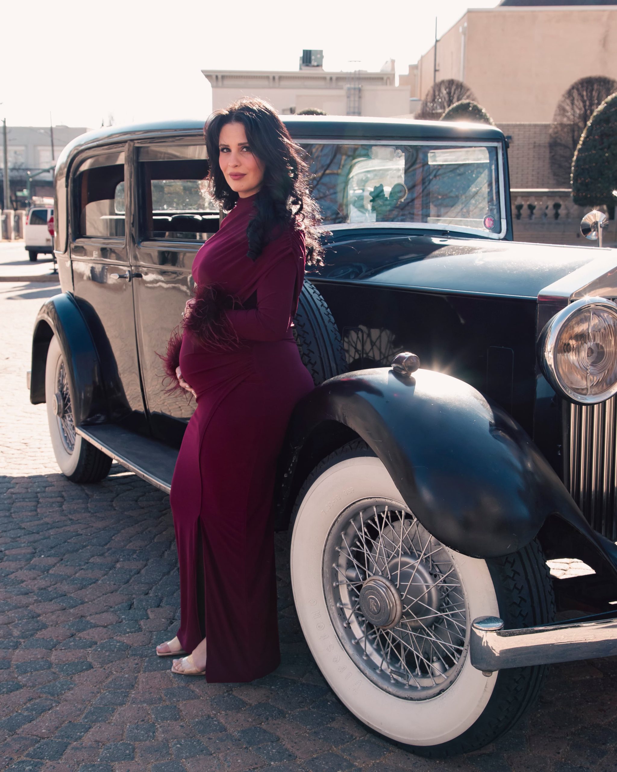 Pregnant woman in a burgundy dress posing beside a vintage dark blue car at Jefferson Hotel in Richmond VA