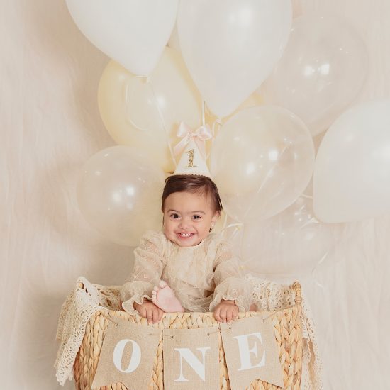 First birthday milestone session featuring a baby girl surrounded by neutral balloons and a ONE prop by Nadia De Sensi Photography.