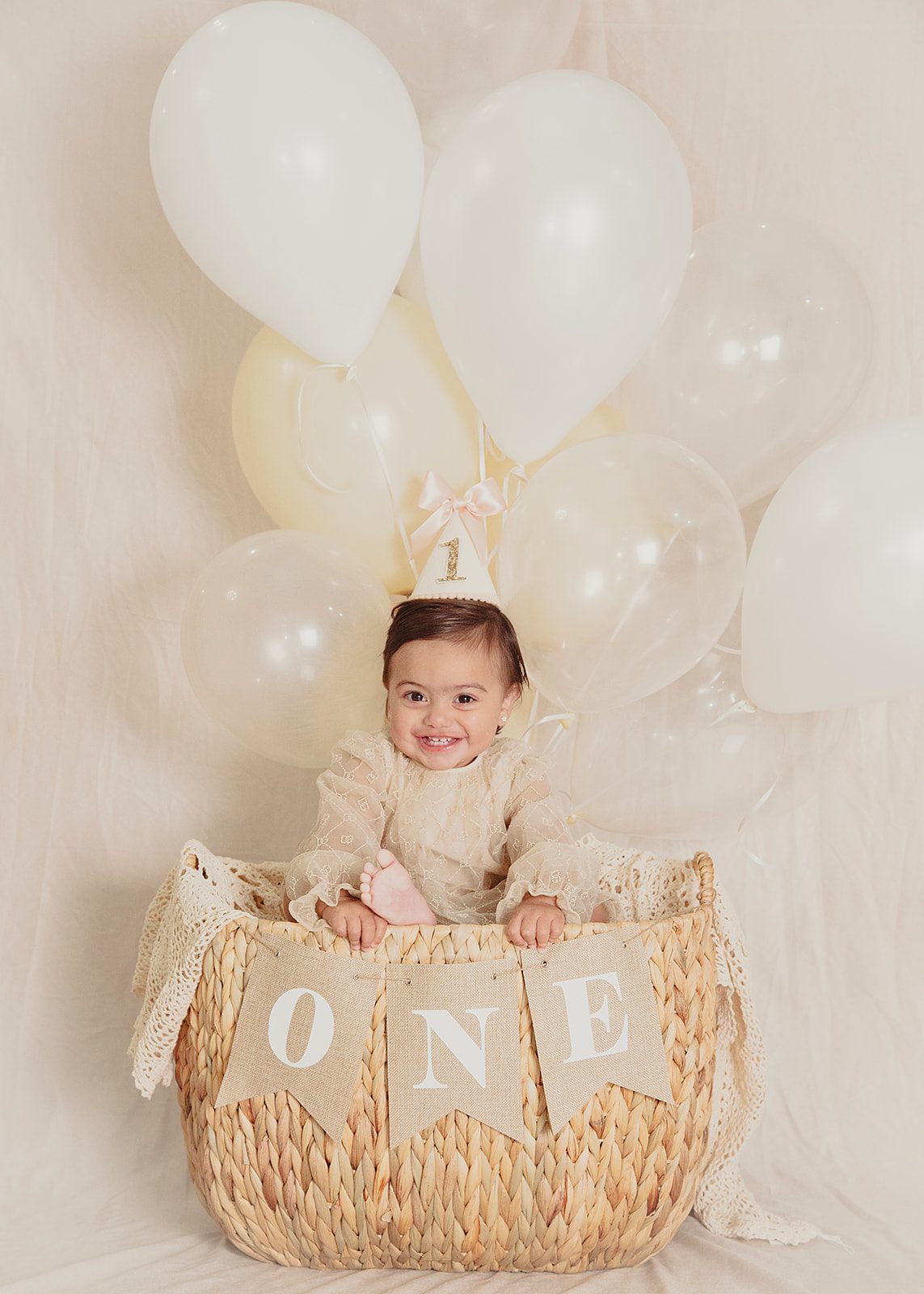 First birthday milestone session featuring a baby girl surrounded by neutral balloons and a ONE prop by Nadia De Sensi Photography.