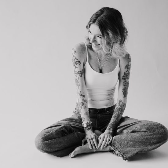 Black and white portrait of a woman with tattooed arms, sitting cross-legged on the floor smiling.